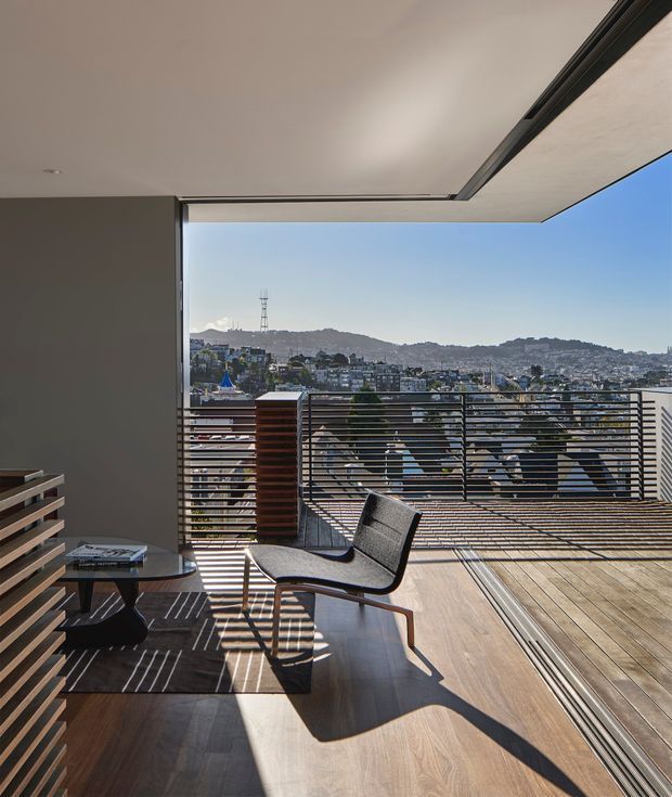 A contemporary indoor-outdoor living area opens onto a spacious timber deck or balcony through fully retractable glazing. The room has dark timber flooring, a neutral feature wall, a low black side table, and a modern upholstered lounge chair. Horizontal metal balustrades frame expansive views across a dense hilly urban landscape. Strong sunlight creates crisp linear shadows across the floor and deck.