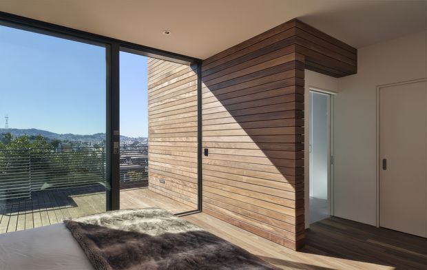 A bright contemporary bedroom with floor-to-ceiling black-framed glazing opening to a balcony or terrace with a city and hillside view. The room features a warm horizontal timber-clad feature wall and projecting timber-lined bulkhead around a doorway, smooth light walls, and dark timber flooring. A low bed or daybed with a dark textured throw occupies the foreground.