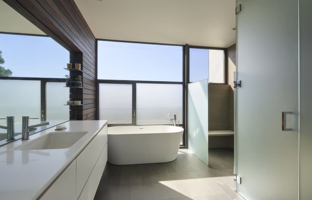 Bright contemporary bathroom with a freestanding white bathtub positioned beside large frosted and clear glazed windows. The room includes a long white vanity with integrated basin and chrome fixtures, dark timber wall cladding with open shelving, dark tiled flooring, and a glass-enclosed shower area. Natural light creates an airy, refined atmosphere.