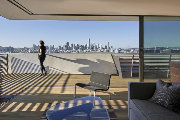 A contemporary rooftop terrace overlooking a dense city skyline and bay, furnished with a dark outdoor lounge chair and sectional sofa. Timber decking, broad glazing, and horizontal slatted privacy screens create strong linear shadows across the terrace in bright afternoon light.