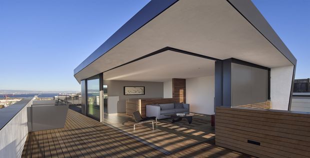 Contemporary rooftop terrace with a covered outdoor living area, expansive glazing, and a minimalist angular roof form. The space includes a lounge seating area, timber decking, dark-framed glass doors, and horizontal timber screening, with an open urban/coastal outlook beyond.