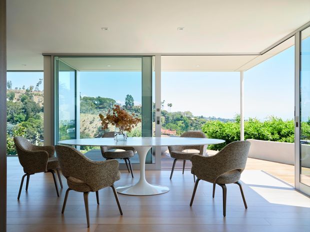 A bright, refined dining area with a round white pedestal table and upholstered dining chairs, positioned beside expansive floor-to-ceiling glazing. The room opens visually to a covered outdoor terrace and a leafy suburban landscape, with pale timber-look flooring, white walls and ceiling, and slim dark-framed sliding glass doors. Natural daylight creates an airy, calm atmosphere.