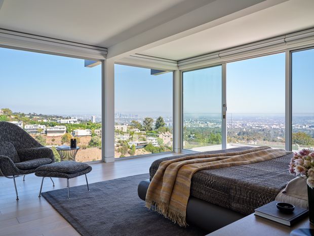 Bright contemporary bedroom with floor-to-ceiling corner glazing and expansive elevated city views. The room features a low upholstered bed, textured grey bedding, a draped brown throw, a dark area rug, and a sculptural lounge chair with ottoman. White walls and ceiling, pale timber flooring, and minimal detailing create an airy, refined interior.