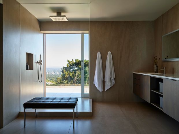 Contemporary bathroom with a walk-in shower, frameless glass opening to a scenic outdoor view, warm timber wall and cabinetry finishes, a floating vanity with white benchtop, and a black upholstered bench. The room has a refined, minimalist feel with integrated shelving and a large glazed opening.