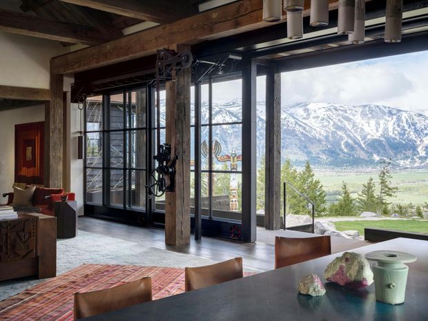 A rustic industrial-style dining and living area with exposed timber beams and posts, dark metal-framed floor-to-ceiling glazing, and a large dining table in the foreground. The room overlooks a dramatic mountain valley, with a sliding barn-style door, sculptural pendant lights, and eclectic furnishings adding character.