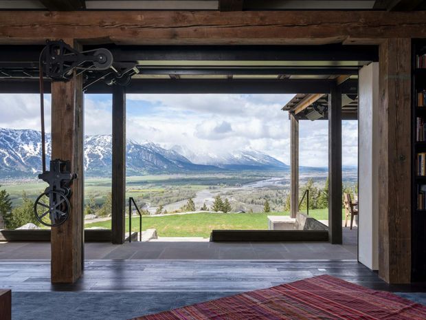 A rustic, dramatic interior opening onto a sweeping alpine mountain and valley view. Heavy reclaimed timber beams and posts frame expansive floor-to-ceiling glazing, with exposed dark metal hardware and a broad dark timber floor. A colourful woven rug occupies the foreground, while the open threshold leads to a terrace and lawn outside.
