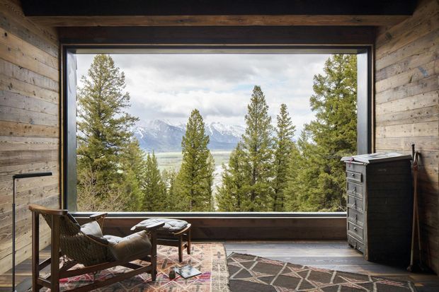 Rustic timber-lined interior lounge or reading nook with exposed dark ceiling beams, a large picture window framing a forested mountain landscape, a leather lounge chair, small side table, patterned rugs, and a dark chest of drawers. The space has a warm cabin-like character and strong connection to the surrounding alpine setting.
