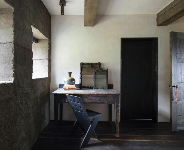 A rustic, dark-toned interior entry or hallway with rough textured plaster walls, deep-set window openings, exposed timber ceiling beams, dark timber flooring, and black-painted timber doors. A distressed wooden console desk with a small decorative vase and mirror is paired with a folding dark chair, creating a moody, heritage-inspired vignette.
