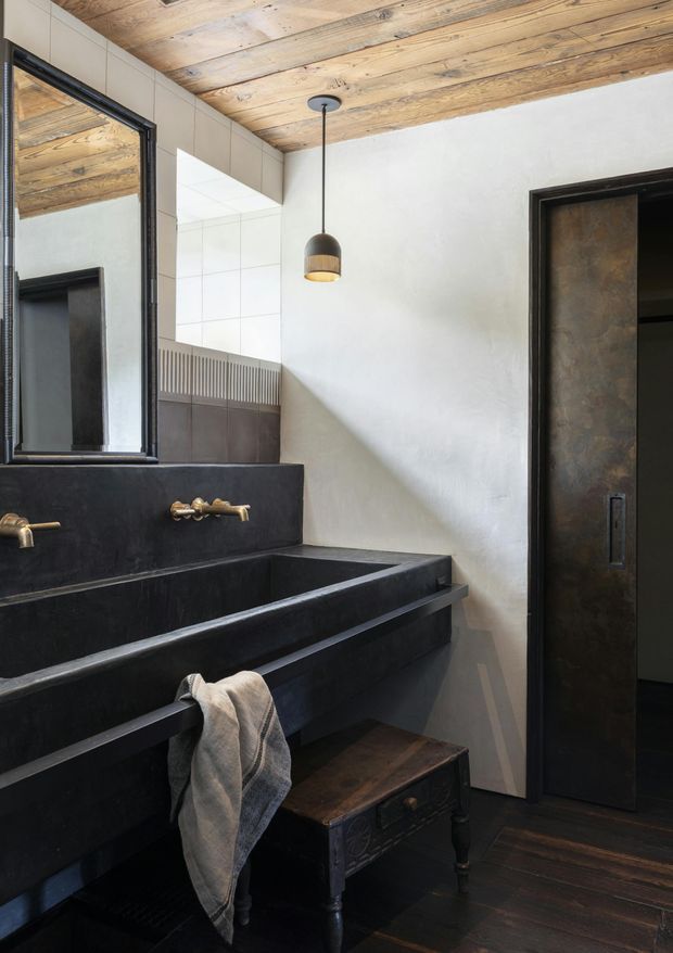 A moody, characterful bathroom with a large black built-in double vanity, exposed brass wall-mounted taps, and a tall black-framed mirror. The room features a warm reclaimed timber plank ceiling, white plaster walls, dark timber flooring, black-framed joinery, and a dark metal or patinated door. A simple pendant light and vintage wooden stool add rustic industrial detail.