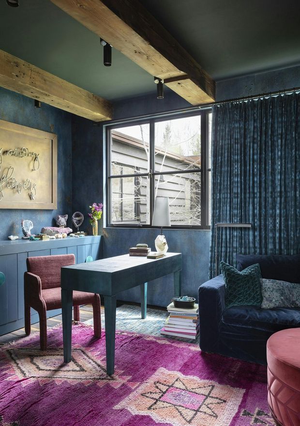 A richly layered, moody home office with deep blue textured walls, dark blue curtains, exposed rough timber ceiling beams, and black track-style spotlights. The room contains a compact dark desk, upholstered burgundy desk chair, dark blue sofa, pink ottoman, blue built-in sideboard, patterned magenta and cream rug, books, and decorative objects. A large steel-framed window provides natural light and looks onto an adjacent structure.
