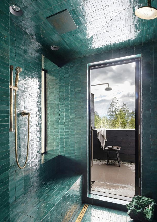 A compact, dramatic shower room clad almost entirely in glossy deep teal-green rectangular tiles, including the walls, ceiling, built-in bench and floor. A wall-mounted brass shower set sits on the left, while a full-height dark-framed glazed door opens to an exterior shower or balcony area with a stool and towel. Skylight-like reflected daylight brightens the ceiling, contrasting with the moody saturated tilework.