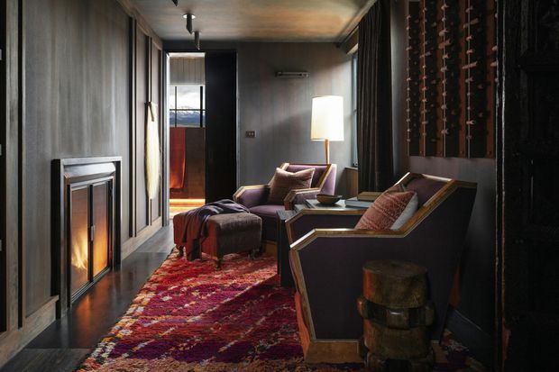 A dark, refined lounge or media room with a linear fireplace, two angular upholstered armchairs, an ottoman, a floor lamp, and a richly patterned red rug. The space features dark vertical wall panelling, dark flooring, a warm metallic-toned ceiling, and a built-in wine rack along the right wall. A glazed opening at the rear brings in limited natural light and views beyond.
