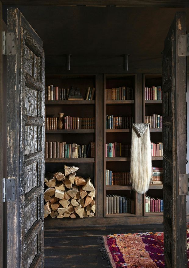 A dark, rustic library or reading nook viewed through heavy weathered timber doors. Floor-to-ceiling dark timber bookcases are filled with books, with a stacked wood store at the base, a hanging woven textile, dark timber flooring, and a colourful patterned rug. The space has a warm, intimate, historic cabin-like atmosphere with exposed aged wood and low lighting.