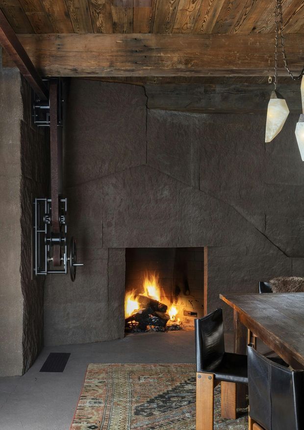 Rustic, moody interior centered on a large open masonry fireplace with a live fire. The room has dark textured plaster or stone-like walls, exposed timber ceiling boards and beam, a timber dining table with black leather chairs, a vintage patterned rug, wall-mounted metal hardware, and pendant lights.