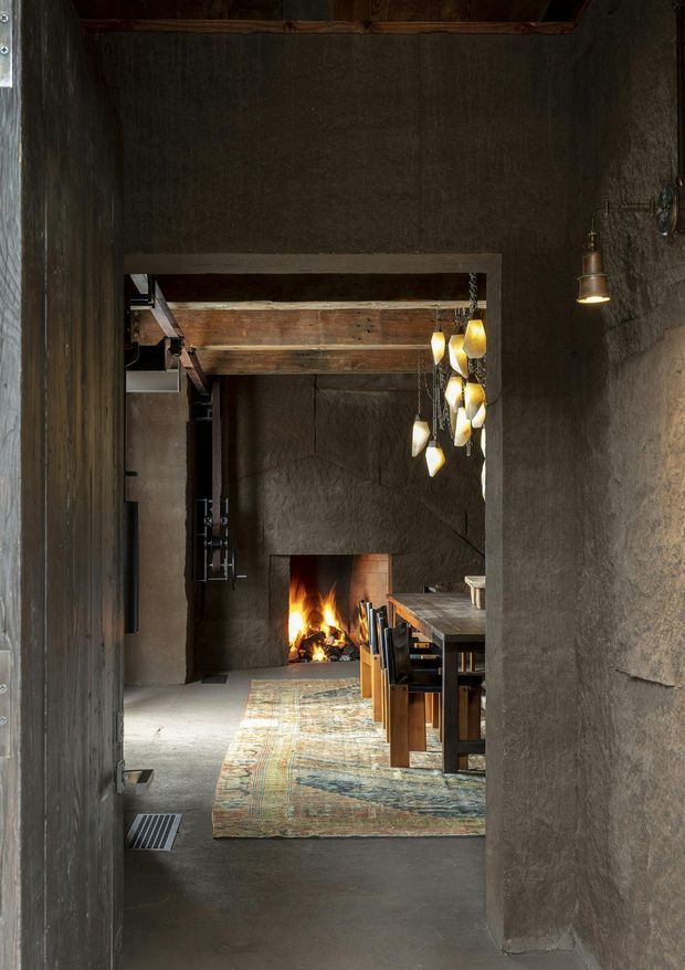 A narrow, moody interior passage looks through to a dining area with a long timber table and chairs, a patterned rug, exposed timber ceiling beams, and a small open fireplace. The walls and floor have dark, rough plaster or concrete-like finishes, with warm pendant and wall lighting creating a rustic, atmospheric setting.