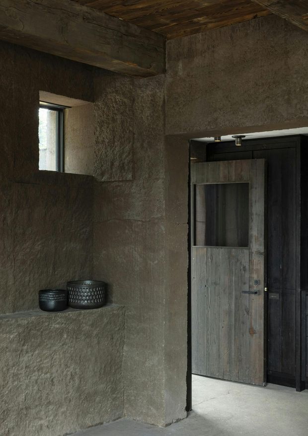 A dim, rustic interior passage or entry area with heavily textured earthen-plaster walls, a rough timber ceiling, a small recessed window, and a built-in ledge holding two dark vessels. A weathered timber door opens into a darker adjoining space, with black fixed joinery or panelling around it. The palette is predominantly deep earthy brown, charcoal, and muted grey, creating a raw, moody atmosphere.