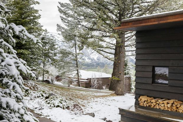 Snow-covered mountain or alpine property landscape viewed beside a dark timber-clad cabin or outbuilding. The building has horizontal dark cladding, warm timber soffit and trim, and a small window, with neatly stacked firewood stored beneath an exterior ledge. Snow-laden evergreen trees, a sloping snowy garden, and distant mountainous terrain are visible.