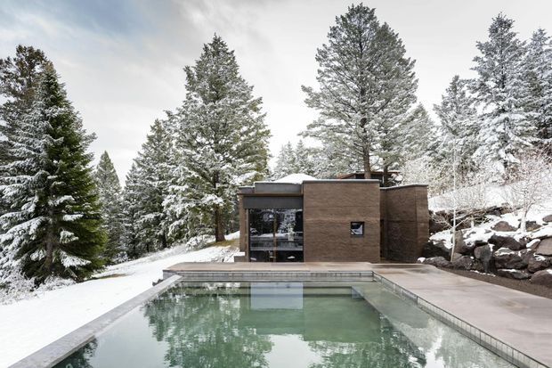 A modern flat-roofed concrete and dark brown-clad pool house or pavilion set within a snow-covered alpine forest. The compact building has extensive glazing, a paved terrace, and a large rectangular outdoor pool in the foreground, with snow-covered rocks and tall coniferous trees surrounding the site.