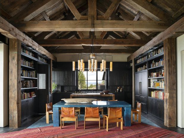 A dramatic, rustic study-library with a central blue stone or painted dining/work table, leather chairs, extensive dark built-in bookshelves, and a compact kitchen or bar area at the rear. The room features exposed heavy timber post-and-beam construction, a pitched roof with exposed rafters, dark timber wall panelling, and a sculptural linear pendant light. A window at the back provides a view beyond, while a patterned red rug adds warmth and contrast.