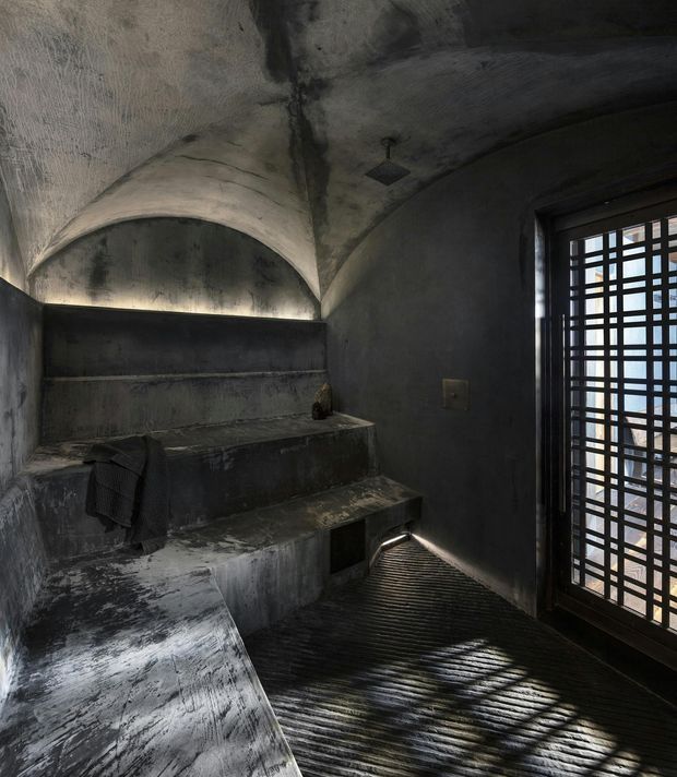 A dark, atmospheric steam-room or sauna interior with heavily textured charcoal concrete surfaces, sculptural vaulted ceilings, and tiered built-in concrete benches. A partially open metal-grid door admits strong linear daylight across the ribbed dark floor, creating dramatic shadows and highlights. The space has a raw, monolithic, cave-like character.