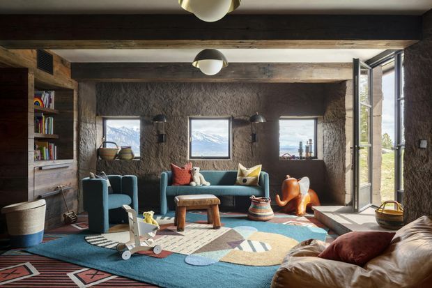 A rustic, cozy living room with rough earthen-textured walls, exposed dark timber beams, built-in timber shelving, and multiple windows overlooking a mountainous landscape. The room contains a teal sofa and armchairs, a tan leather sofa, colourful patterned rugs, a low timber coffee table, pendant lights, and playful decorative furnishings. A glazed external door is open to the right, bringing in bright natural light.