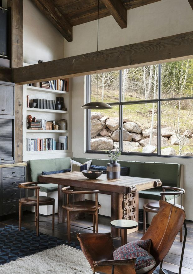 A rustic, refined dining area with a large timber dining table, built-in upholstered bench seating, sculptural wooden dining chairs, and a leather lounge chair in the foreground. Exposed dark timber beams and posts frame the ceiling and upper wall, while a large black-framed window overlooks a rocky wooded landscape. Built-in shelving displays books and ceramics beside dark timber cabinetry. The room combines warm natural materials with muted green upholstery and dramatic structural detailing.