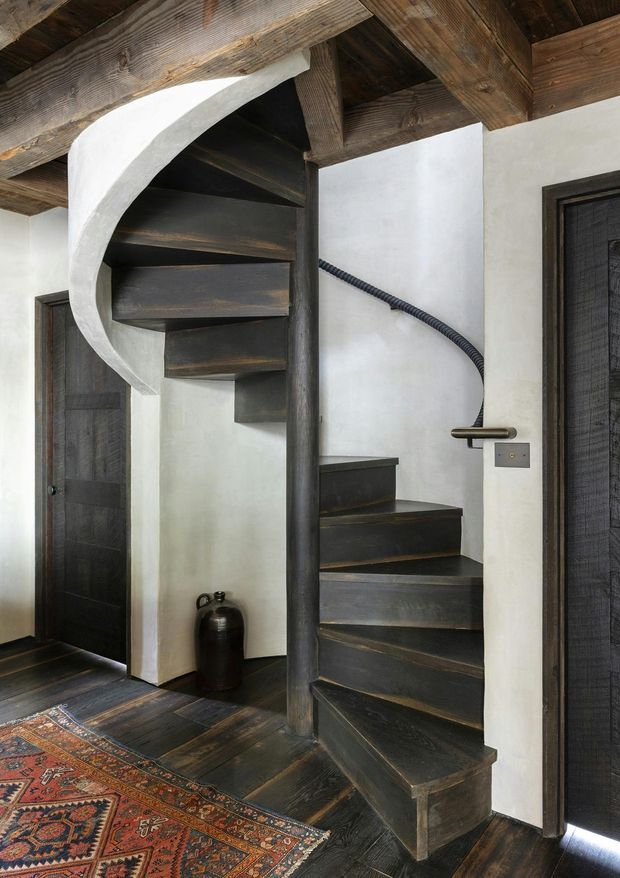 Interior view of a compact spiral staircase with dark timber treads, central timber newel, and a curved white plaster stair stringer. The staircase sits within a rustic, richly textured interior with exposed dark timber ceiling beams, dark timber floors and doors, white plaster walls, a brass-toned handrail, and a patterned Persian-style rug.