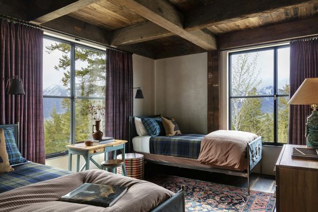 A cozy rustic bedroom with two single beds, dark timber ceiling beams and exposed timber ceiling boards, large black-framed windows, and deep plum curtains. The room includes layered plaid and textured bedding, a patterned area rug, a small timber side table, bedside lamps, and a timber dresser. Mountain and tree views are visible through the windows.