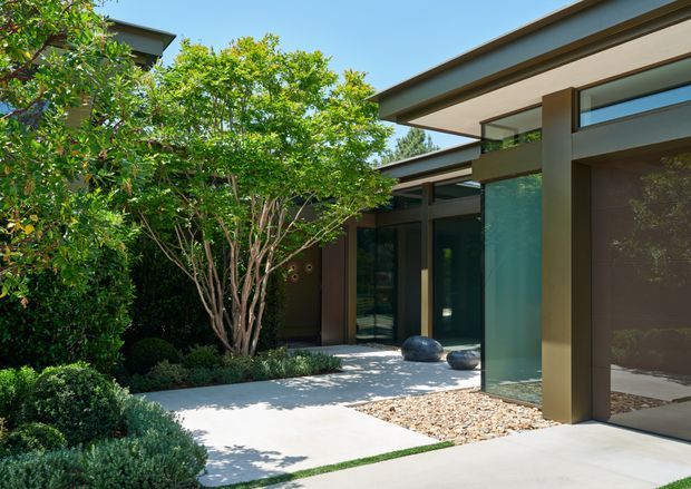 A modern single-storey residential exterior enclosing a landscaped courtyard. The building has flat roof planes with deep overhangs, dark bronze or brown structural frames, large floor-to-ceiling glazing, and a minimalist pale concrete path and paving. Dense clipped greenery, a small ornamental tree, sculpted shrubs, and a gravel bed soften the geometric architecture.