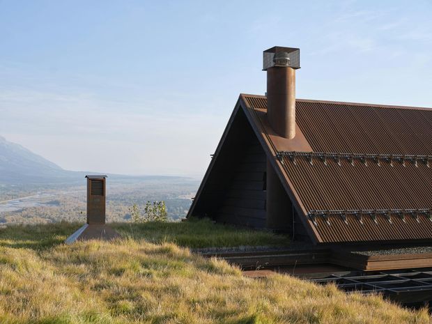 Exterior view of a dark timber-clad, steeply pitched gable-roof structure with corrugated metal roofing and a prominent cylindrical metal chimney. The building appears integrated into a grassy roof or landscape, with expansive mountain and valley scenery in the background.