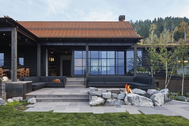 Exterior view of a dark timber-clad residence with a broad rust-coloured pitched metal roof, large black-framed glazed openings, and a covered outdoor entertaining area. Stone-paved terraces and steps lead to a landscaped lawn, with a built-in outdoor fire feature formed by large natural boulders in the foreground. Forested hills are visible beyond the property.