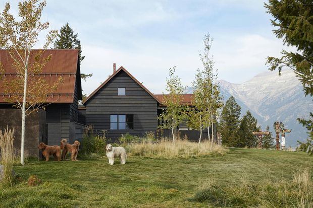 A dark timber-clad mountain house or lodge with steep pitched gable roofs and rust-red metal roofing, set within a grassy landscaped site. Young deciduous trees, mature conifers, and a dramatic mountain backdrop create a rustic alpine setting. Several dogs are present in the foreground, and a decorative signpost stands near the property edge.