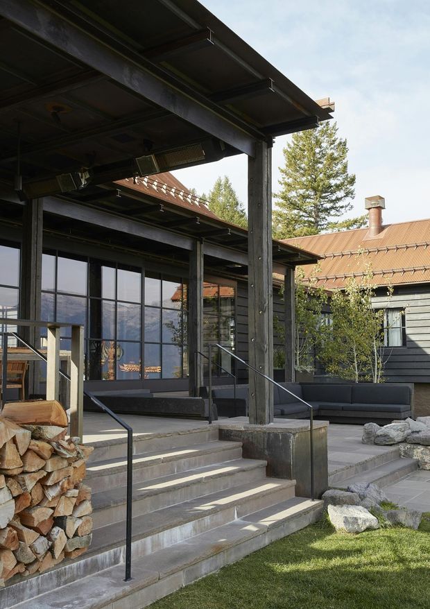 Exterior view of a substantial timber-clad building with a covered entry terrace, exposed dark structural posts and beams, broad steel-edged roof canopies, large black-framed glazed doors, and steps leading into an outdoor seating area. The building has a corrugated metal pitched roof with a chimney, dark horizontal cladding, timber decking and paved terraces. Stacked firewood, large rocks, trees and distant mountains are visible around the entry.