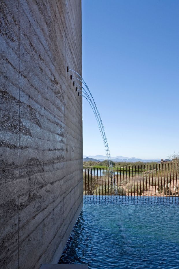 Vertical architectural exterior detail showing a rectangular swimming pool beside a tall textured concrete block wall. Water streams arc from small wall outlets into the pool, with a dark metal safety fence beyond. A dry, open landscape and distant mountain range are visible under a clear blue sky.