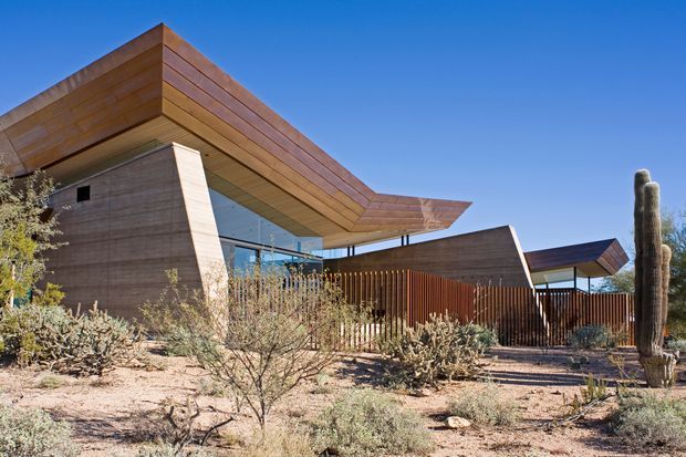 Exterior architectural photograph of a contemporary single-storey building set within an arid desert landscape. The structure features angular cantilevered roof planes, warm horizontal timber-toned cladding, extensive glazing, and a screened courtyard or entry area with vertical timber slats. Desert shrubs and tall columnar cacti surround the building under a clear blue sky.