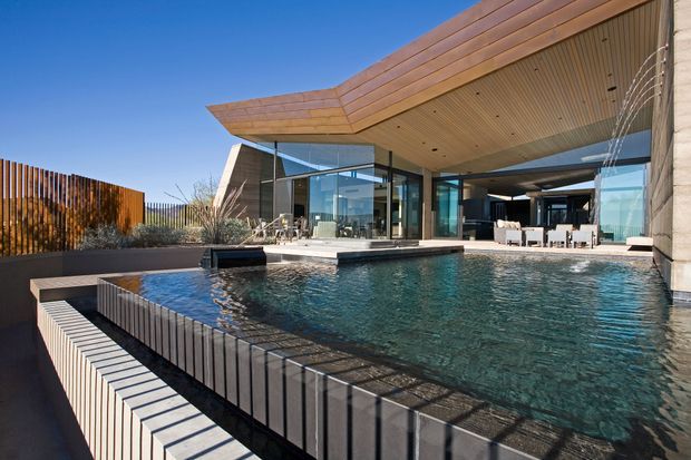 Contemporary luxury residence viewed from an outdoor pool courtyard. A large rectangular swimming pool with a raised dark-tiled edge dominates the foreground, with water features along the right side. Behind it is a low, angular pavilion with extensive floor-to-ceiling glazing, warm timber soffits and roof cladding, exposed concrete walls, and an open-plan living or entertaining area. Vertical timber screening and minimalist landscaping frame the property under a clear blue sky.