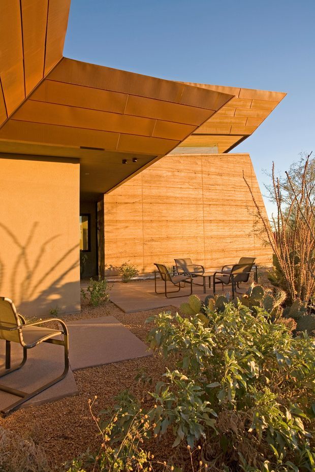 Exterior architectural photograph of a contemporary low-rise building with a dramatic cantilevered, angular roof canopy clad in warm copper-toned metal panels. The facade combines smooth light plaster with a large textured earthen or concrete wall, opening onto a paved outdoor seating courtyard with modern metal chairs and tables. Desert-style landscaping with gravel, shrubs and cacti surrounds the building, under a clear blue sky.