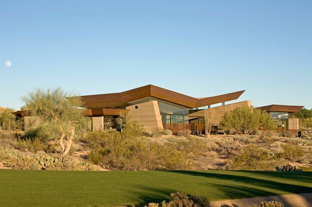A low-slung contemporary residence set in an arid desert landscape, with broad cantilevered roof planes, warm wood-toned soffits, sandy masonry walls, extensive glazing, and sparse native planting. A manicured green lawn occupies the foreground, contrasting with the rocky, scrub-covered site. The building has a strong horizontal composition and appears largely single-storey.