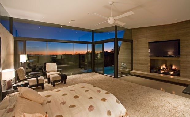 Spacious, warmly lit bedroom with floor-to-ceiling corner glazing overlooking the dusk landscape, a low-profile bed, lounge chairs, wall-mounted television, and a built-in fireplace set into a textured timber feature wall. The room has a soft carpeted floor, a ceiling fan, and a contemporary resort-like feel.