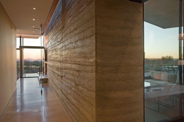 A refined contemporary interior corridor dominated by a thick, rough-textured rammed-earth feature wall. The polished concrete floor and ceiling-mounted recessed downlights lead toward full-height glazing and an outdoor terrace, while a lounge area with seating is visible through the glass on the right. Warm sunset light and distant hills are visible beyond the glazing.