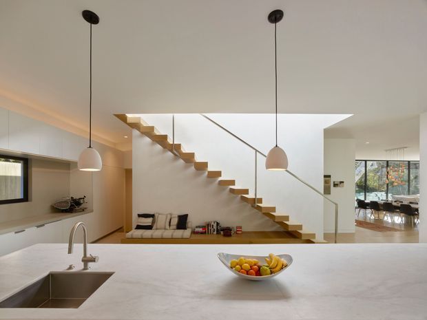 Open-plan contemporary kitchen viewed across a large pale stone island benchtop, with an undermount sink and gooseneck tap in the foreground. The space includes streamlined light cabinetry, pendant lights, recessed ceiling lighting, and a floating timber staircase with a slim glass balustrade. A living area is visible beneath and beside the stair, with a dining area beyond.