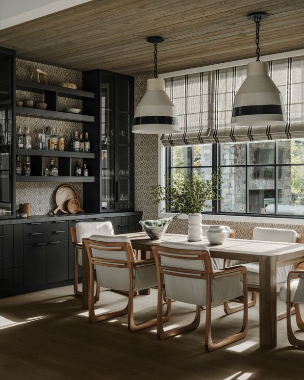 A sophisticated dining area combined with a built-in bar and display cabinetry. The room features black cabinetry, patterned wallpaper, a timber-planked ceiling, dark timber flooring, a large timber dining table, upholstered wood-framed dining chairs, and oversized pendant lights. Full-height windows with woven patterned Roman blinds bring in natural light and reveal a stone exterior beyond.