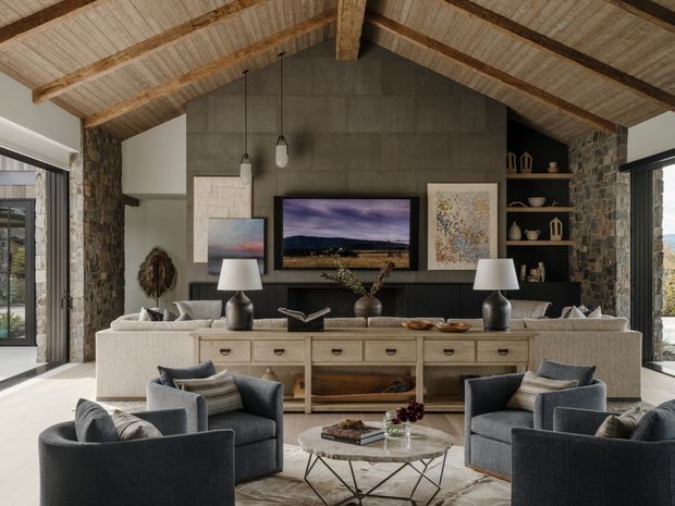 A refined living room with a dramatic vaulted ceiling, exposed timber rafters and beams, and rough stone-clad walls. A large dark fireplace or feature wall anchors the room, flanked by built-in open shelving and artwork. Two sofas face a central seating arrangement with dark upholstered armchairs and a round coffee table, while a long timber console sits behind the seating. Large glazed openings bring in abundant daylight and views of the surrounding landscape.