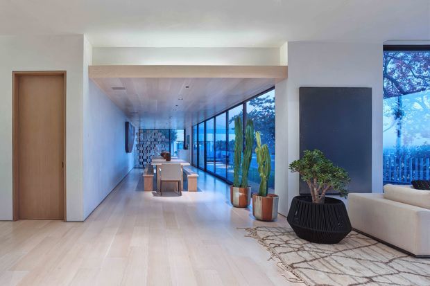 Open-plan contemporary interior with a light timber floor and ceiling, white walls, a compact dining area and kitchen beyond, and a living area beside expansive floor-to-ceiling glazing. A sculptural black planter, potted cacti, and a patterned rug add contrast, while a timber-framed opening and exposed structural beam define the space.