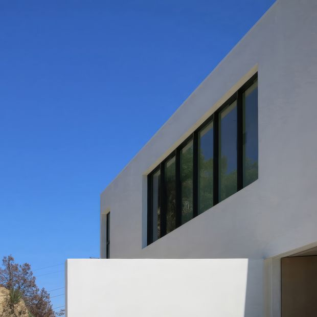 Minimalist two-storey building exterior with a smooth white rendered facade, flat roofline, and a long strip of tall dark-framed windows. A projecting white privacy wall partially obscures the lower level, while clear blue sky and sparse trees form the background.