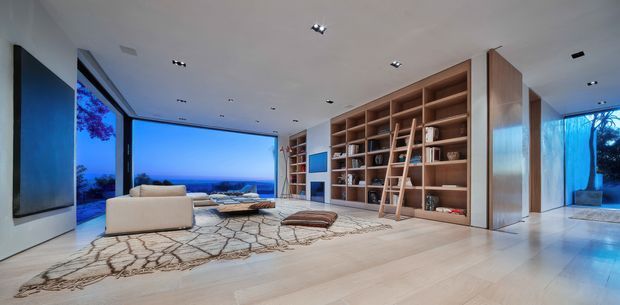 A spacious, contemporary living room with expansive floor-to-ceiling glazing overlooking a blue coastal or ocean-like horizon. The room features pale timber flooring, a large neutral patterned rug, low cream seating, recessed ceiling lights, and a dramatic full-height built-in timber shelving wall with a leaning ladder. A large wall-mounted dark panel is visible on the left, while glazed openings connect to an exterior terrace or pool area on the right.