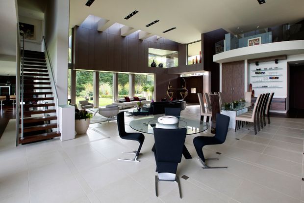 A spacious, open-plan contemporary interior combining a dining area, living area, kitchen, and stairway. The foreground features a round glass dining table with sculptural dark upholstered chairs, while a second dining or kitchen counter area sits to the right. Floor-to-ceiling glazing opens the living space toward a landscaped garden. Dark timber wall panelling and cabinetry contrast with pale tiled flooring, white ceilings, and extensive integrated lighting. A floating open-riser staircase is visible on the left, and built-in shelving and display joinery line the right wall.