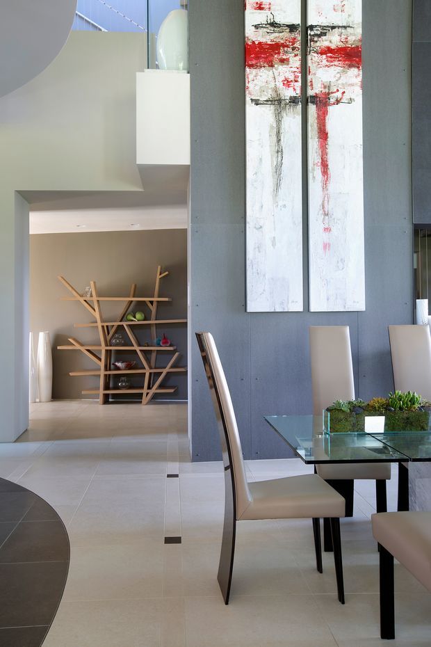 A refined contemporary dining area with a glass-top rectangular dining table, upholstered cream dining chairs, and a long low planter centerpiece. The room features a tall charcoal-grey textured wall with two large abstract red, black, and white artworks, pale tiled flooring with dark geometric inlays, and a sculptural white pendant light. An open passage leads to a display alcove with distinctive branch-like timber shelving and decorative objects.