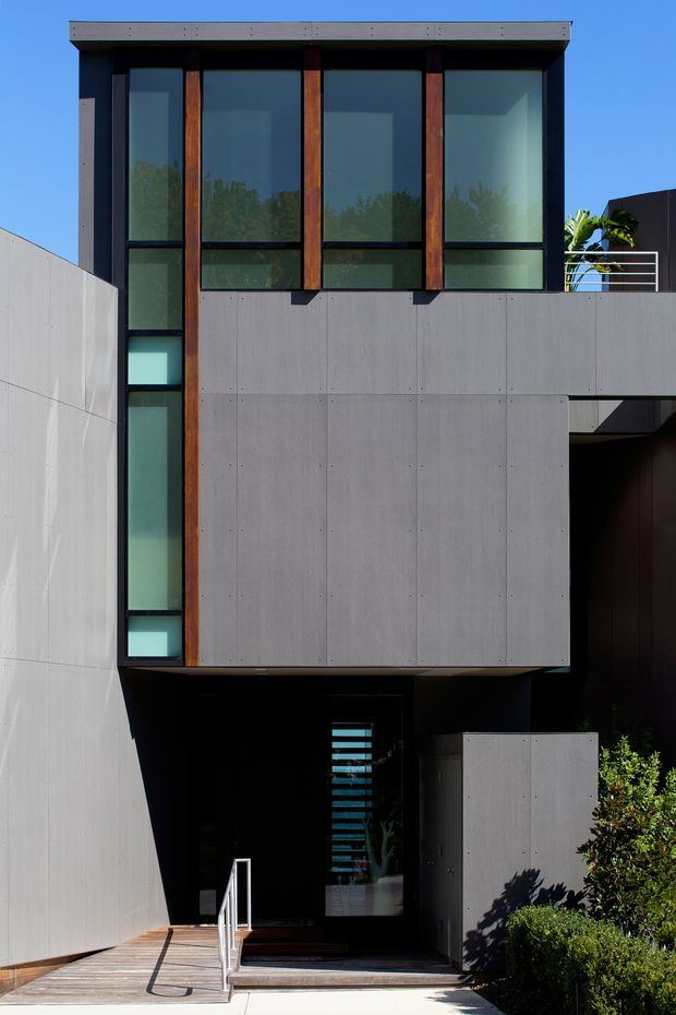 Photograph of a contemporary multi-storey residential facade with a strong rectilinear composition. The building combines smooth grey concrete or fibre-cement panels, dark window framing, extensive floor-to-ceiling glazing, and vertical warm timber accents. A recessed entry is reached by a small ramp and stair, while a terrace with a horizontal metal balustrade is visible at upper right. The facade is shown in bright daylight with strong shadows and landscaped planting near the entrance.