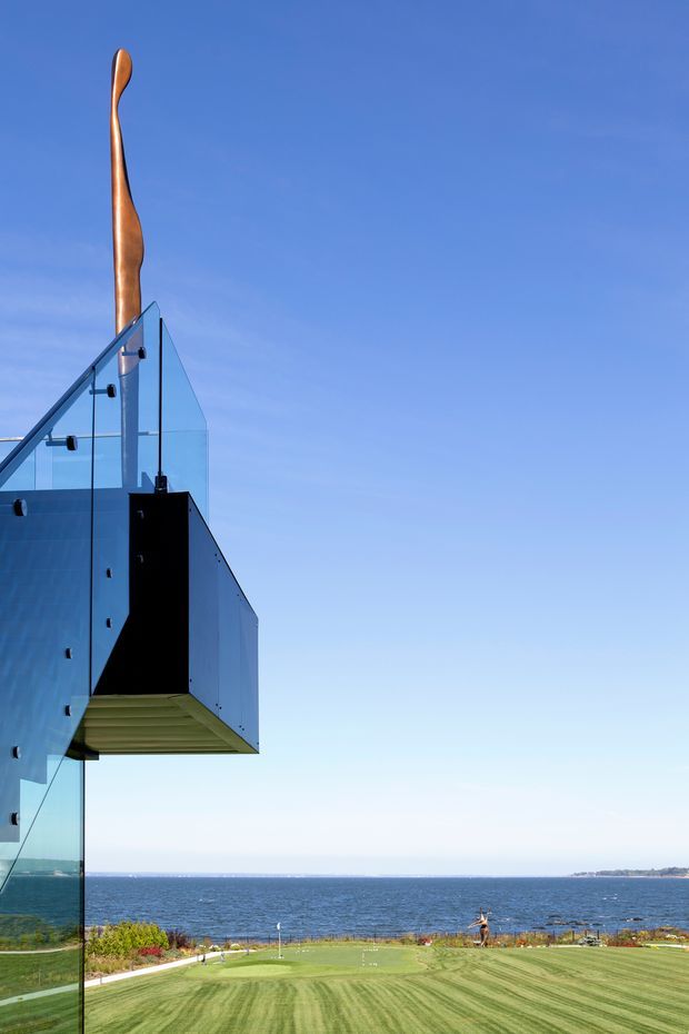 Exterior architectural view of a contemporary glass-clad structure projecting over a landscaped coastal property. The image features transparent glass balustrades, a dark projecting wall or volume, and a distinctive tall bronze sculptural element rising above the roofline. A broad manicured lawn and ocean horizon are visible beyond, under a clear blue sky.